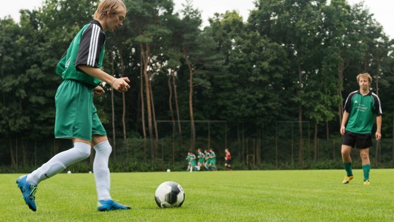 Zwei junge männliche Fußballspieler in grün-schwarzen Trikots kicken sich auf einem Outdoor-Fußballplatz einen Fußball zu. 