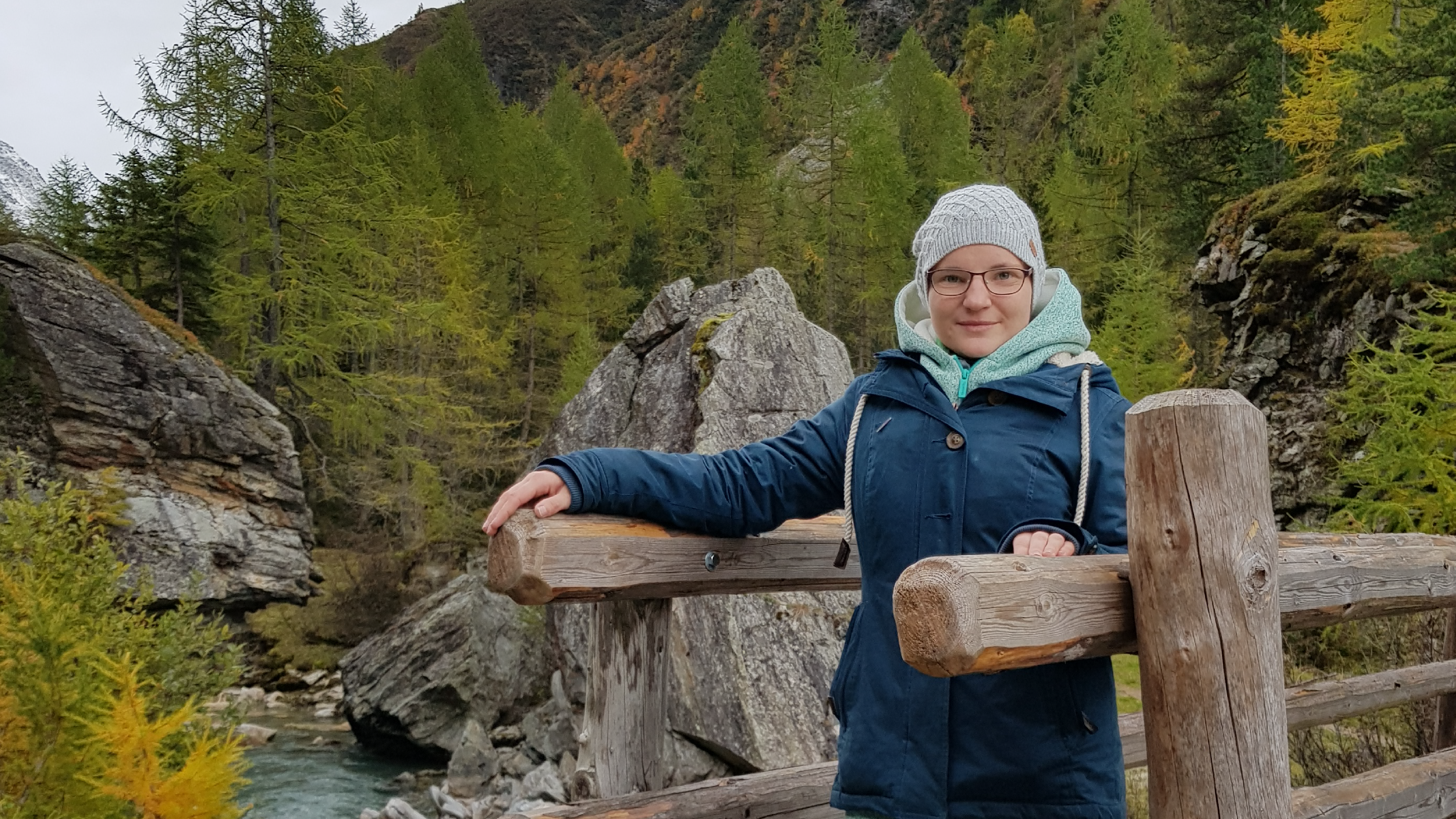 Eine Frau steht auf einer Holzbrücke über einem Fluss. Sie trägt eine Mütze und einen blauen Anorak. 