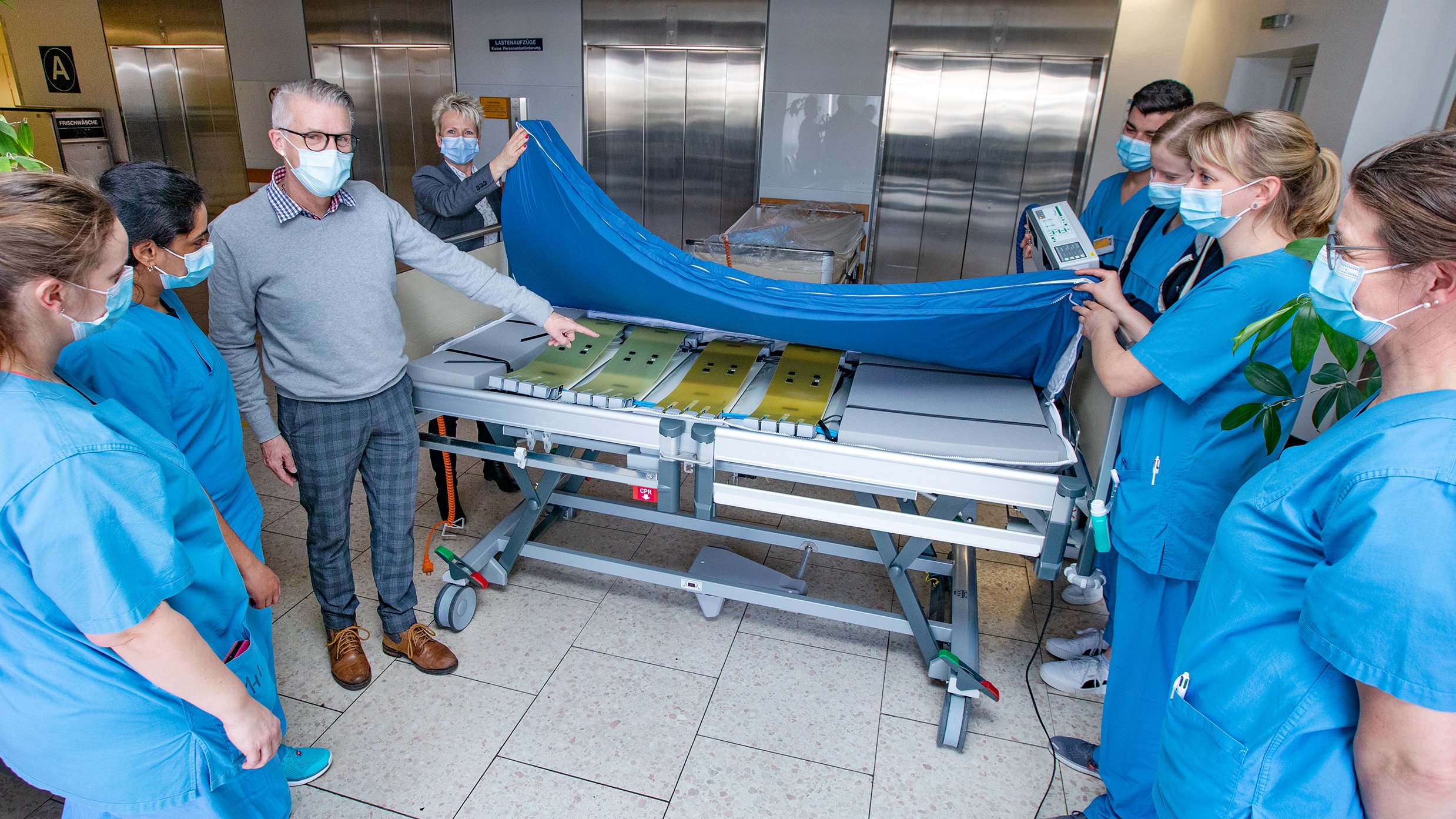 Mehrere Pflegekräfte heben die neu angeschaffte Matratze an. Der Mitarbeiter des Matratzenherstellers zeigt auf die komplexe Technik darunter, die die automatische Mobilisation ermöglicht. 