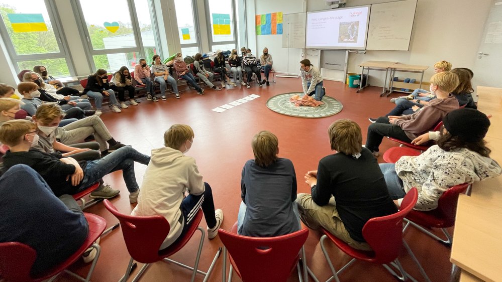 Schüler:innen sitzen im Kreis und in der Mitte erklärt ein Studierender die Grundlagen der Wiederbelebung. 