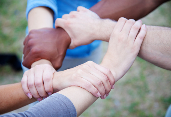 Fünf Hände von Menschen unterschiedlicher Hautfarbe umschlingen jeweils ein anderes Handgelenk und bilden somit einen Kreis.