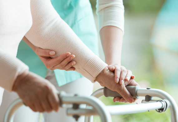 Hände und Arme sind zu sehen. Das Bild lässt erahnen, dass es sich um eine ältere Patientin handeln könnte, die sich an einem Rollator festhält und dabei von einer Pflegekraft unterstützt wird.