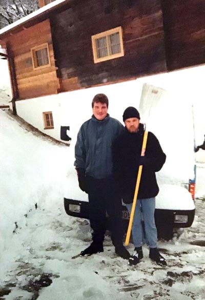 Christian steht als junger Mann mit einer weiteren Person vor einem Haus im Schnee. Copyright: privat