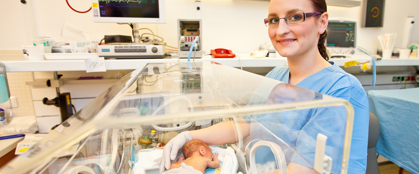 Ein zu früh geborenes Kind liegt in einem Inkubator, neben dem einen Kinderkrankenpflegerin steht. 