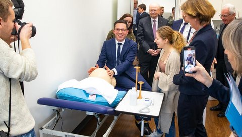 Falko Mohrs sits on a lounger and holds his hands on a dummy stomach. Press representatives, midwives and MHH President Michael Manns stand around him.