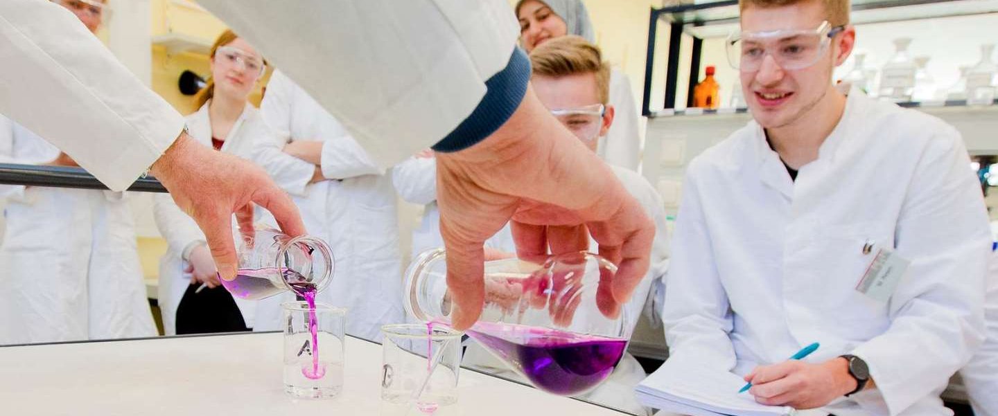 Medizin-Studierende mit Schutzbrillen sitzen vor einem Tisch, auf dem jemand zwei farbige Flüssigkeiten in jeweils ein Glas schüttet. 