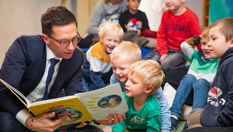 Wissenschaftsminister Falko Mohrs sitzt mit den MHH-Campuskinder auf dem Boden und hält ein Kinderbuch in den Händen.