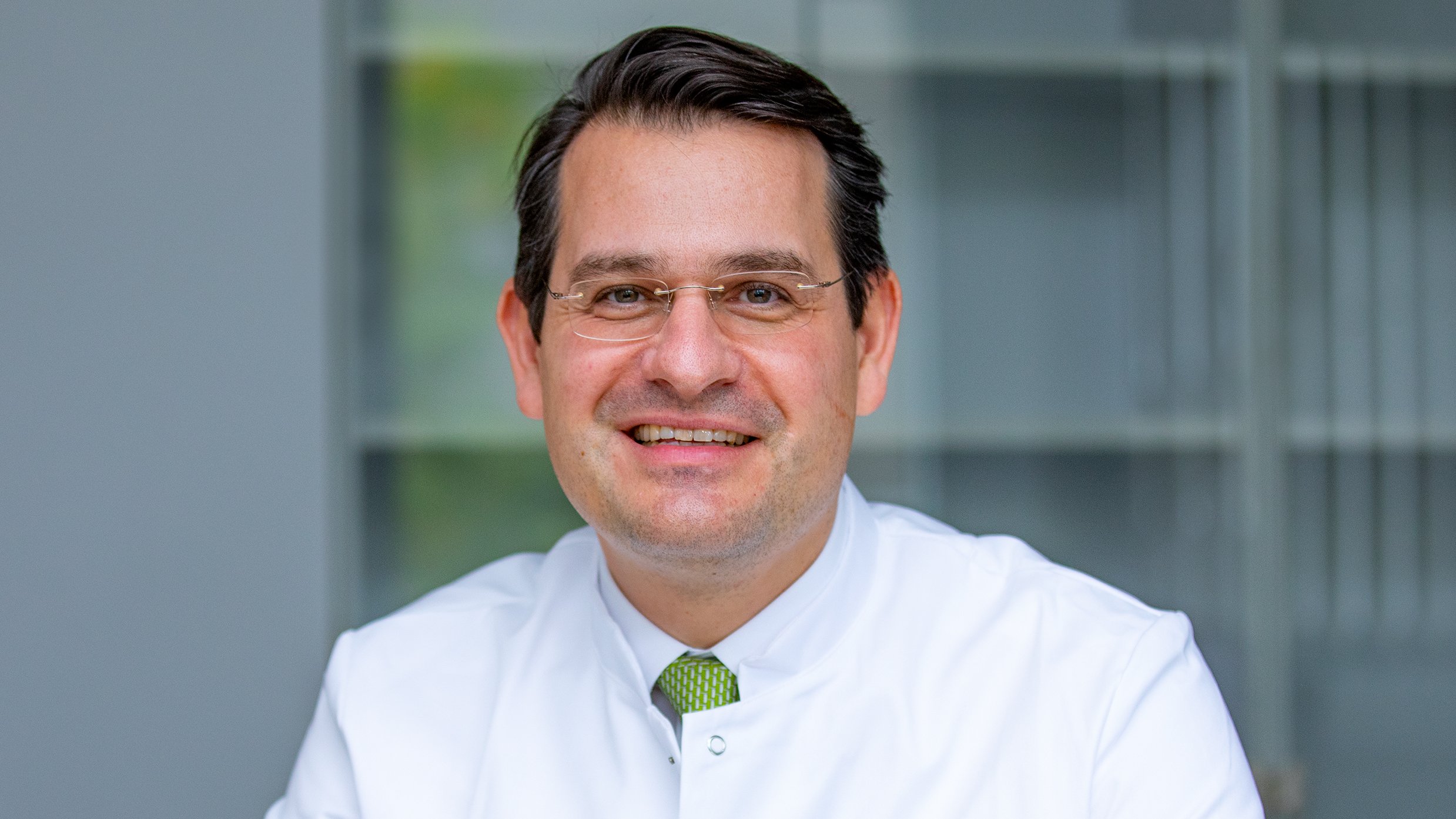 Copyright: MHH/Karin Kaiser Professor Schmelzle sitzt an einem Tisch, trägt Kittel und eine grüne Kravatte und lächelt freundlich in die Kamera, Copyright: MHH/Karin Kaiser