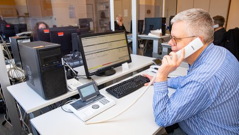 Ein Mann sitzt an einem Schreibtisch mit PC-Monitor und hält einen Telefonhörer am Ohr. 
