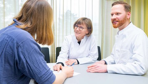 Professorin Trebst und Dr. Hümmert, beide im Arztkittel, sitzen an einem Tisch und unterhalten sich mit einer Patientin, die ihnen gegenübersitzt.