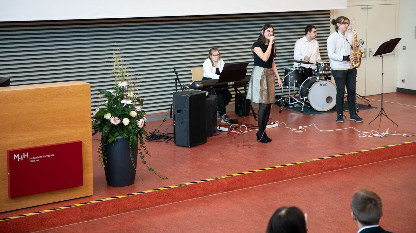 Eine Sängerin steht mit drei anderen Musikern auf dem Podium eines Hörsaals und singt ins Mikrofon. 