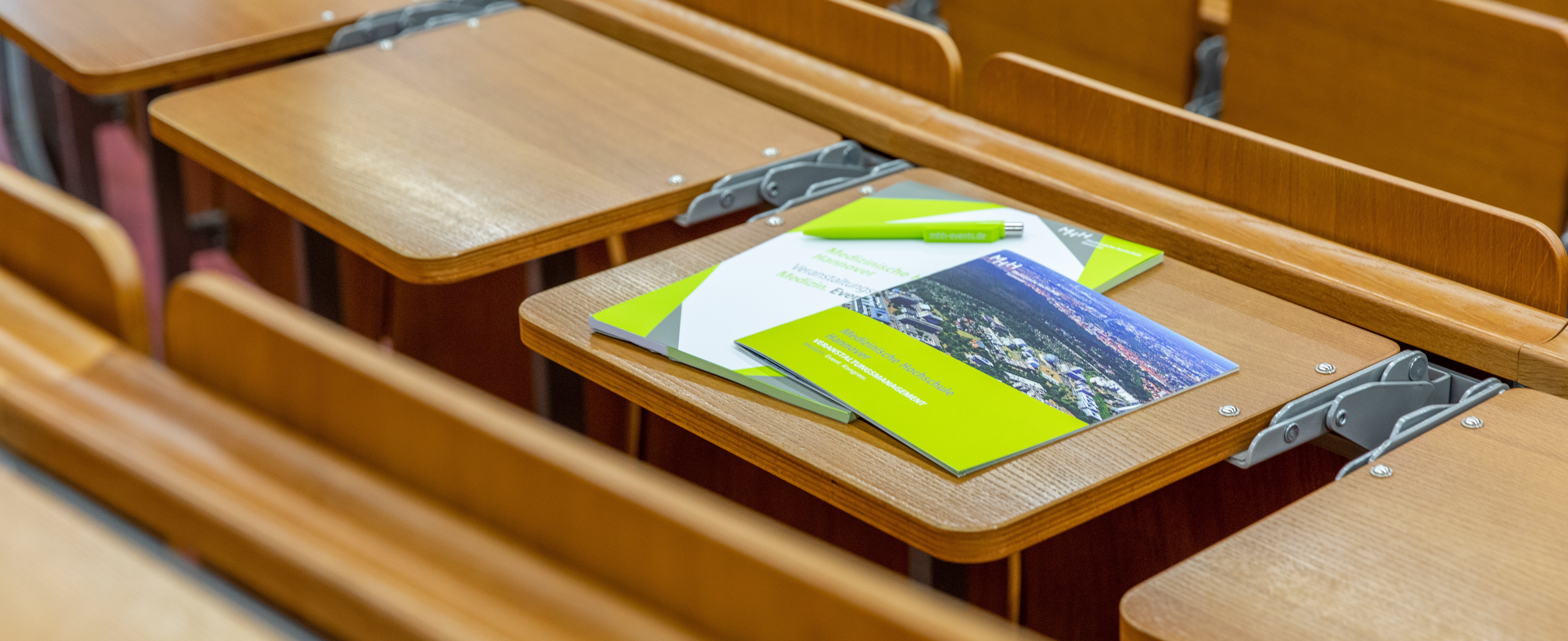 eine Broschüre, Block und Kugelschreiber liegen auf dem Tisch in einem Hörsaal