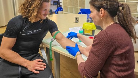 Eine Frau und ein Mann sitzen sich an einem Tisch. Die Frau nimmt dem Mann Blut ab.
