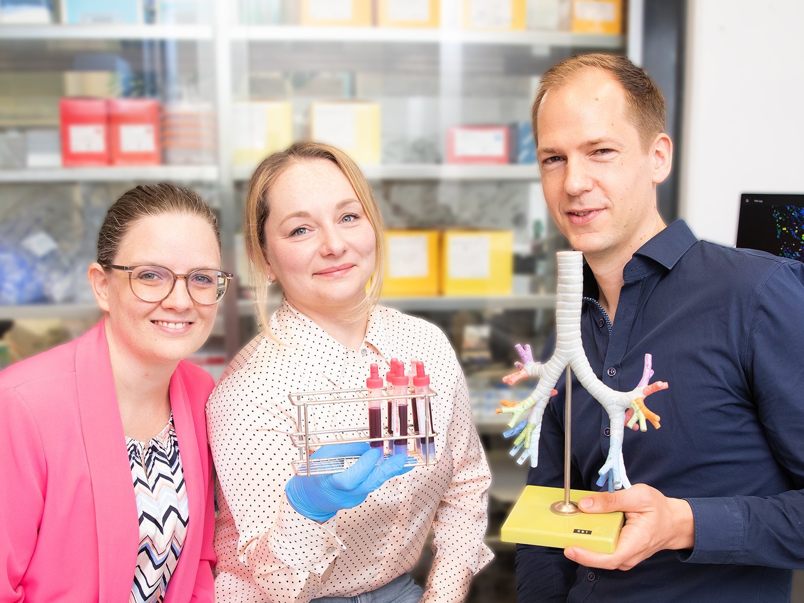 Eine Naturwissenschaftlerin, eine Ärztin und ein Arzt stehen in einem Labor und halten ein Lungenmodell und Blutproben in den Händen.