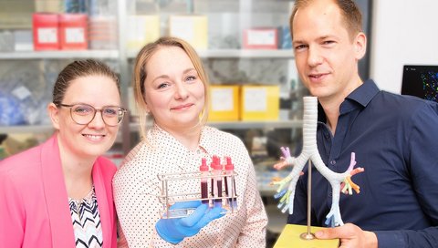 Eine Naturwissenschaftlerin, eine Ärztin und ein Arzt stehen in einem Labor und halten ein Lungenmodell und Blutproben in den Händen.