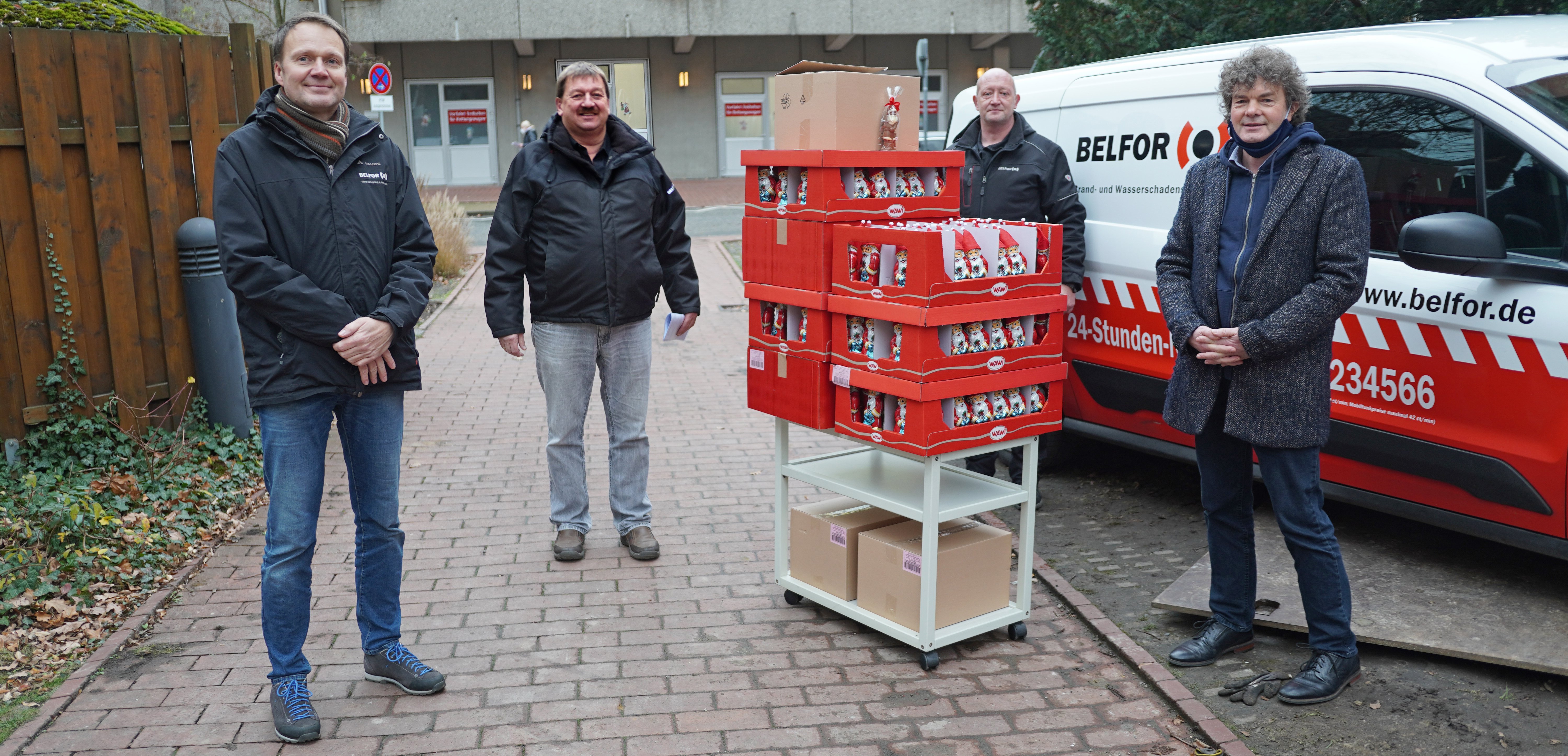 Drei Belfor-Mitarbeiter stehen gemeinsam mit Dr. Schenke vor einem Lieferwagen des Unternehmens. Im Vordergrund ist ein kleiner Transportwagen mit sieben Papppaletten voller Schoko-Weihnachtsmänner zu sehen.