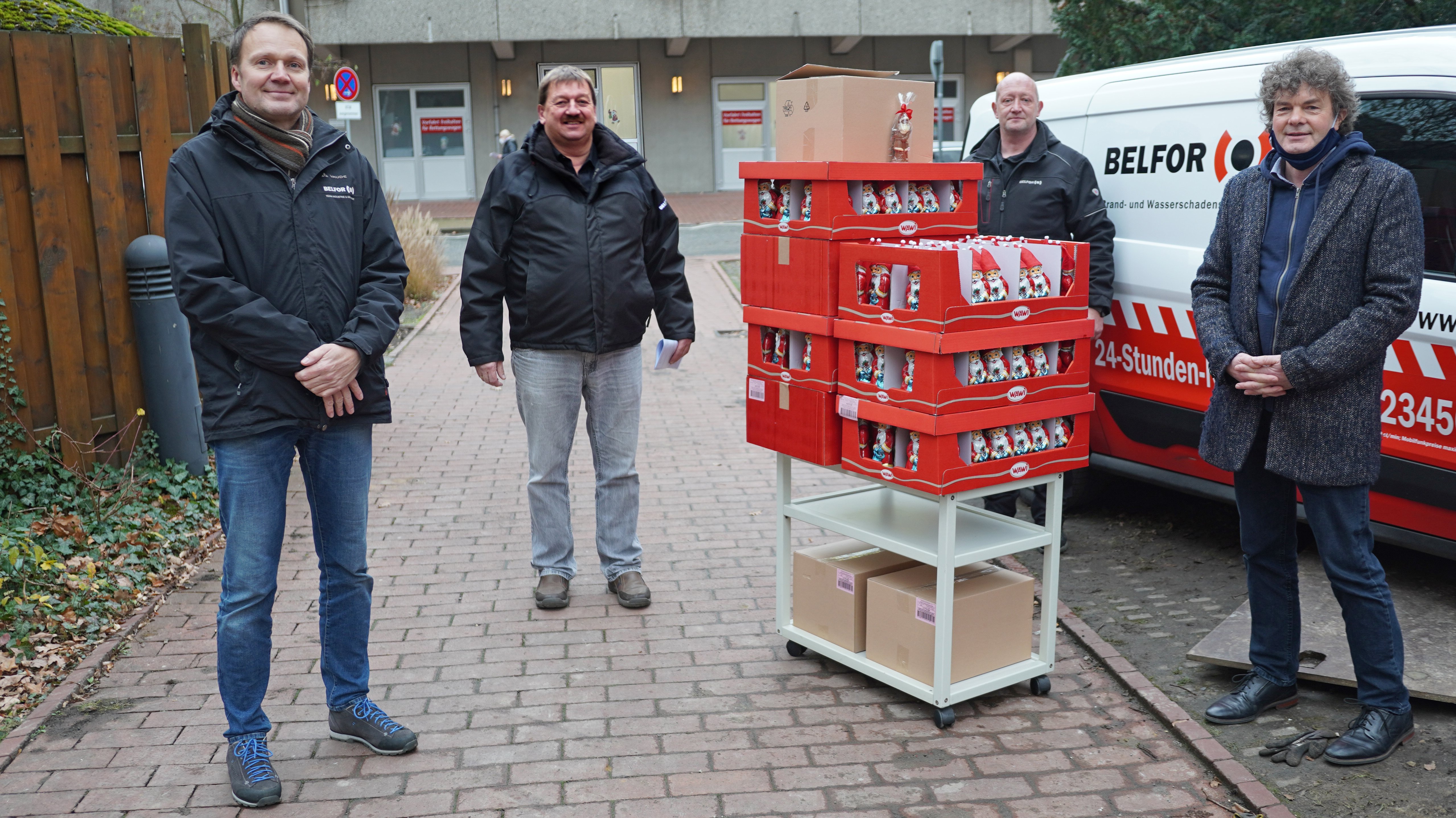 Drei Belfor-Mitarbeiter stehen gemeinsam mit Dr. Schenke vor einem Lieferwagen des Unternehmens. Im Vordergrund ist ein kleiner Transportwagen mit sieben Papppaletten voller Schoko-Weihnachtsmänner zu sehen.