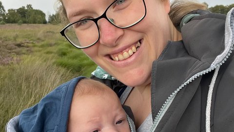Eine Frau trägt in einer Bauchbinde ein kleines Baby. Sie lächelt in die Kamera. Im Hintergrund ist eine grüne Wiese zu sehen. 