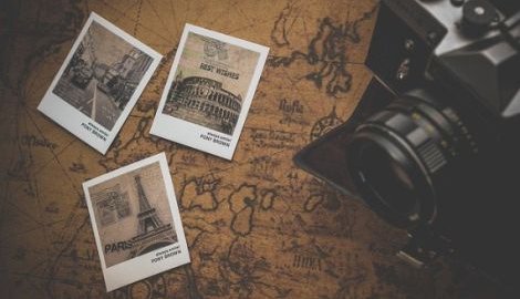 Auf einer alten, vergilbten Weltkarte liegen 3 Polaraidfotos und ein Fotoapparat. Auf einem Polaroid ist der Eifelturm zu sehen, auf einem das Kolloseum und auf dem dritten eine Straße in London mit einem Taxi und einem Bus.