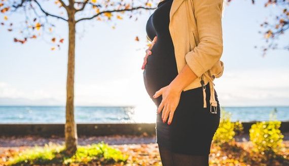 Eine schwangere Frau steht vor einem Gewässer. Davor stehen auf der linken Seite ein Baum und Büsche. Die Frau steht im Vordergrund und hat die rechte Hand zwischen Brust und Bauchansatz gelegt und die linke Hand an der linken Leiste. Die Sonne scheint auf das Gewässer.