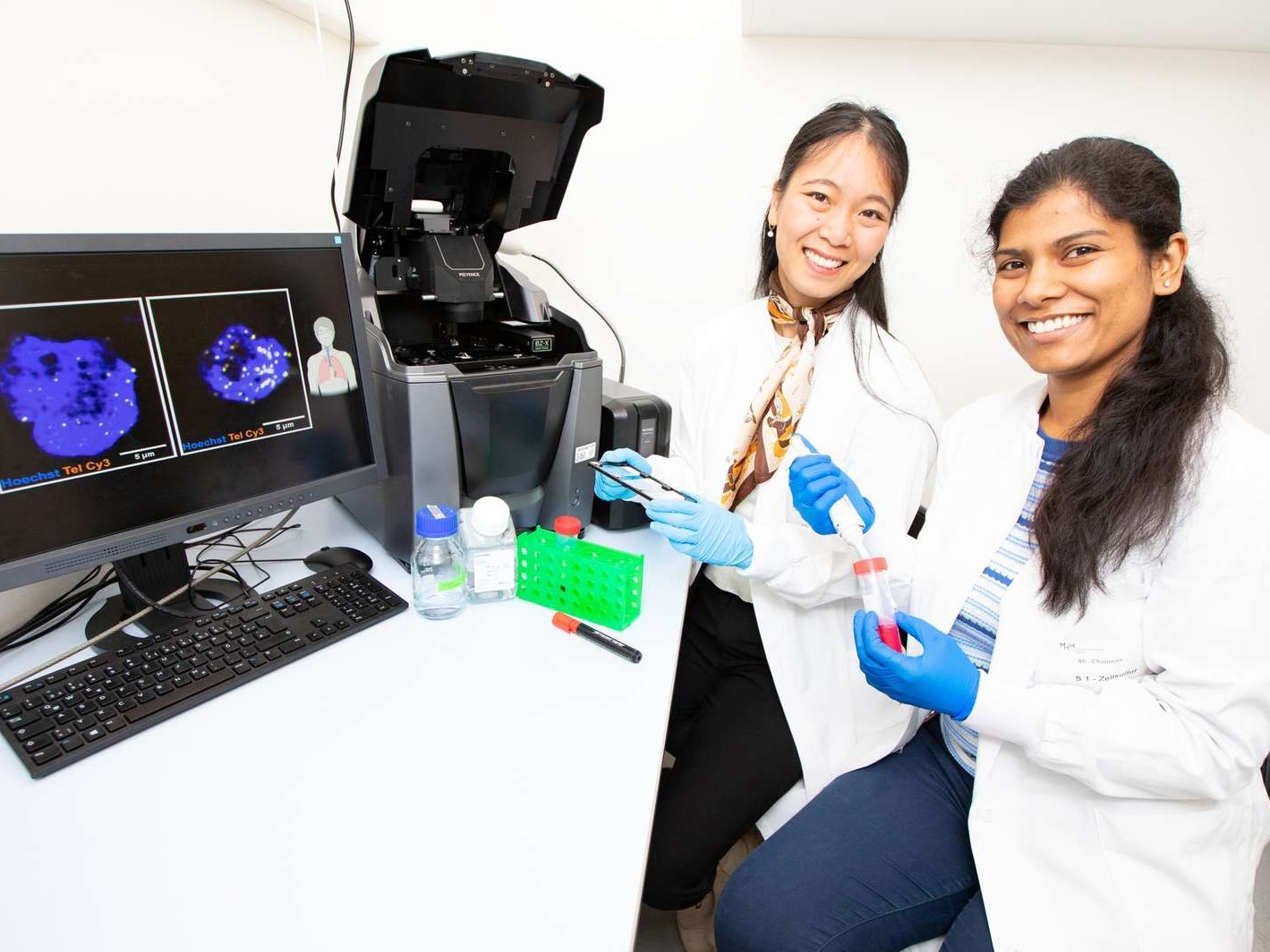 Two women in white coats sit in front of a fluorescence microscope.