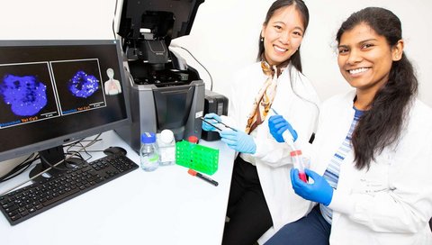 Two women in white coats sit in front of a fluorescence microscope.