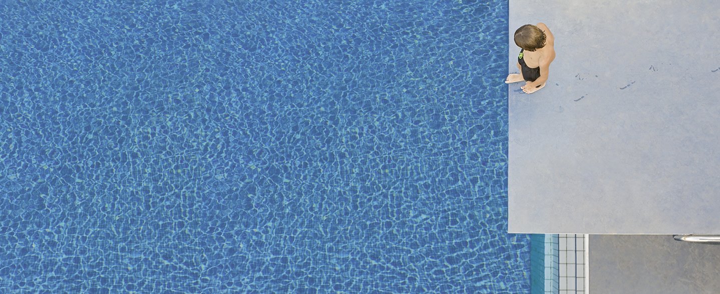 Perspektive von oben: Ein Junge steht auf einem Sprungbrett und blickt auf das Schwimmbecken unter ihm. 