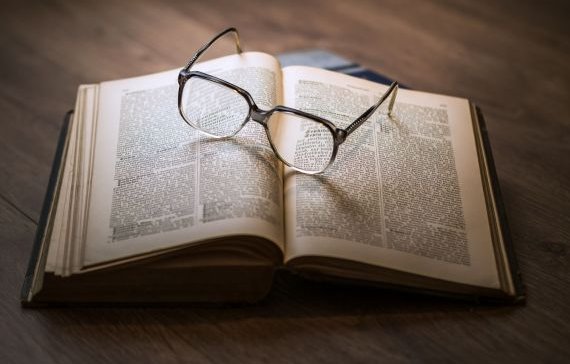 Ein aufgeschlagenes Buch liegt auf einer Holzplatte. Auf dem Buch liegt eine Brille mit einem dicken Rand.