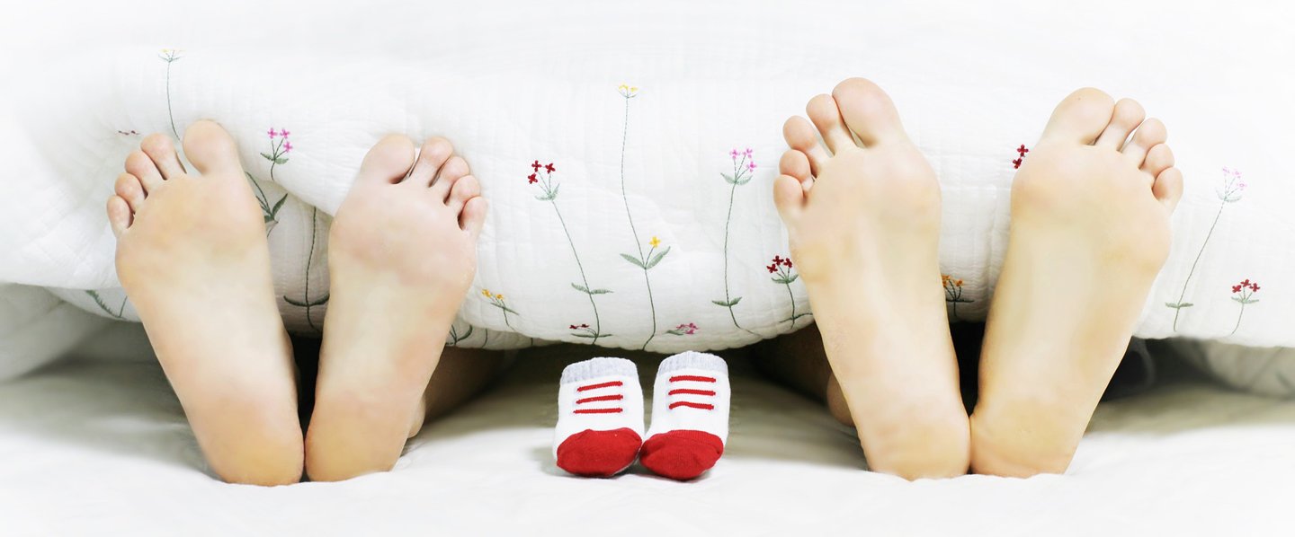 Foto bei pexels Babyschühchen zwischen 2 Fußpaaren lugen unter der Bettdecke hervor