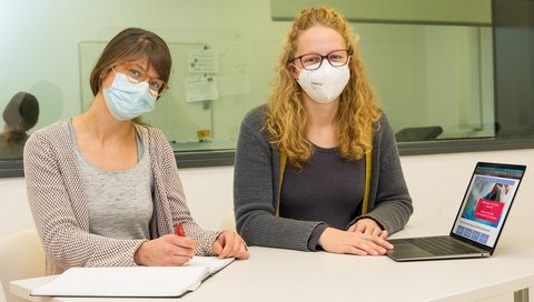 Jacqueline Niewolik (links) und Marie Mikuteit vom ErgoLoCo-Team sitzen mit Mundschutz neben einander an einem Tisch mit aufgeklappten Laptop und einem Heft. 