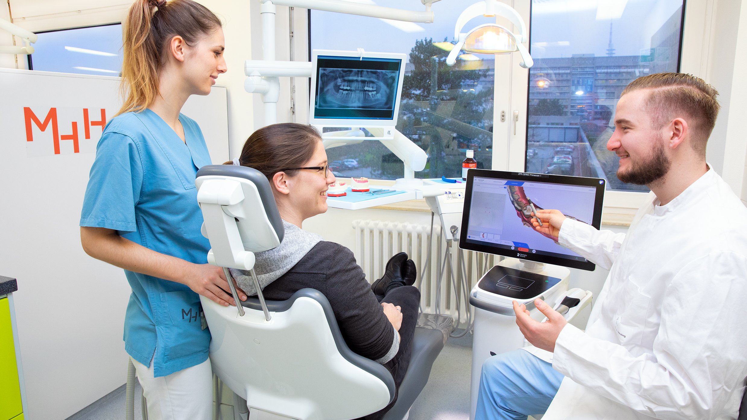 Studierende in blau-weißer Klinikkleidung steht um den Behandlungsstuhl herum, auf dem ein Patient sitzt. Beide blicken zum Zahnarzt, der in weißem Kittel gekleidet auf der rechten Seite sitzt und auf einem Gerät ein digitales Bild erklärt.