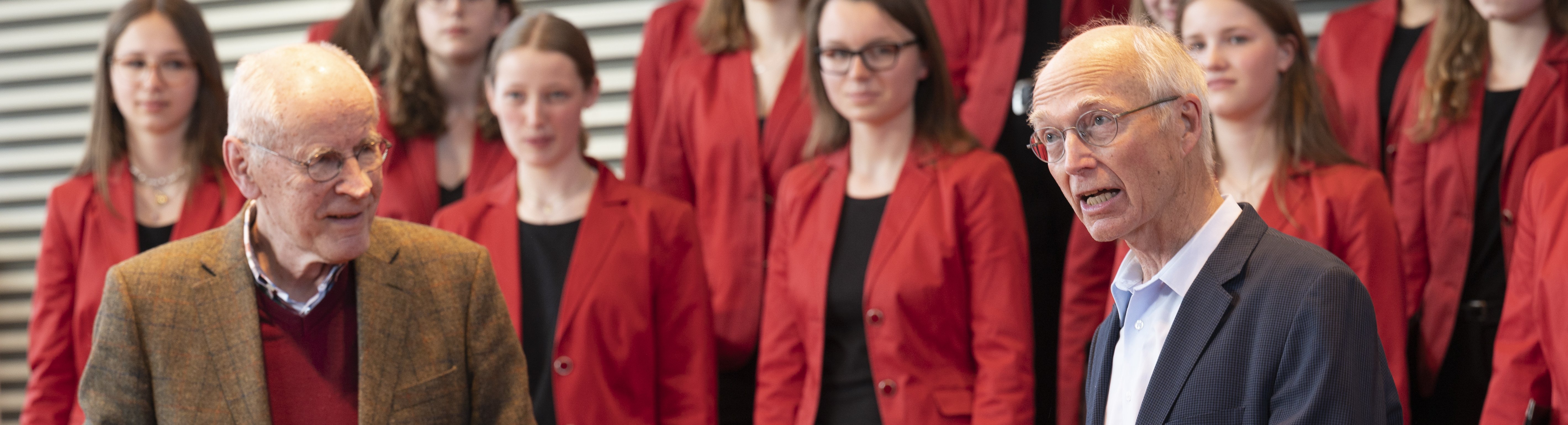 Die Professoren stehen gemeinsam im Vordergrund, hinten ist undeutlich der Mädchenchor zu erkennen.