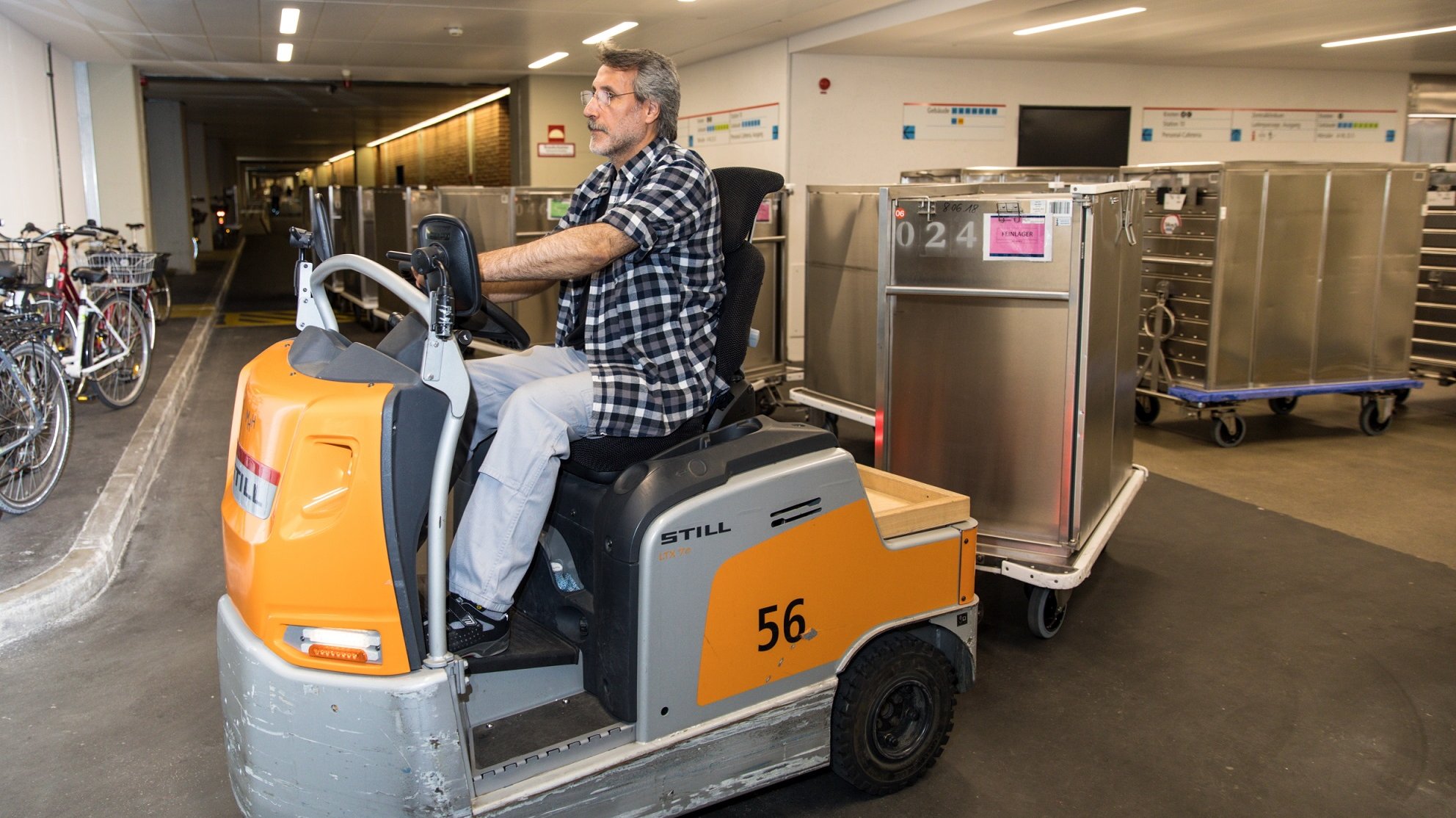 Mitarbeiter des Transport auf einer Elektrolok mit mehreren angekoppelten Transportwagen fährt auf einem Flur der MHH um die Kurve und im Hintergund stehen weitere Transportwagen zur Abholung bereit