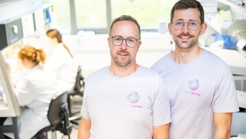 Zwei Männer stehen nebeneinander im Labor und lächeln freundlich in die Kamera. Im Hintergrund sind Personen zu sehen, die an einer Laborbank arbeiten.