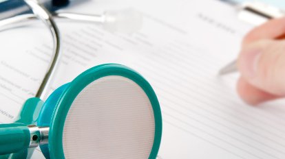 Copyright: pabijan - stock.adobe.com/Institut für Allgemeinmedizin und Palliativmedizin/MHH Auf einem Tisch liegende Kladde, auf die ein Zettel geheftet ist. Auf Zettel und Kladde liegt wiederum ein Stethoskop. Eine Hand hält einen Stift und füllt den Zettel aus.