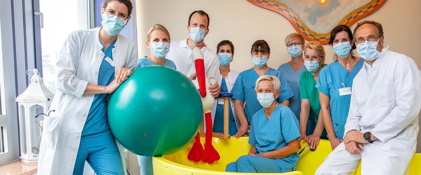 Geburtshilfeteam mit Coronaschutzmasken umstellt eine gelbe Gebärbadewanne 