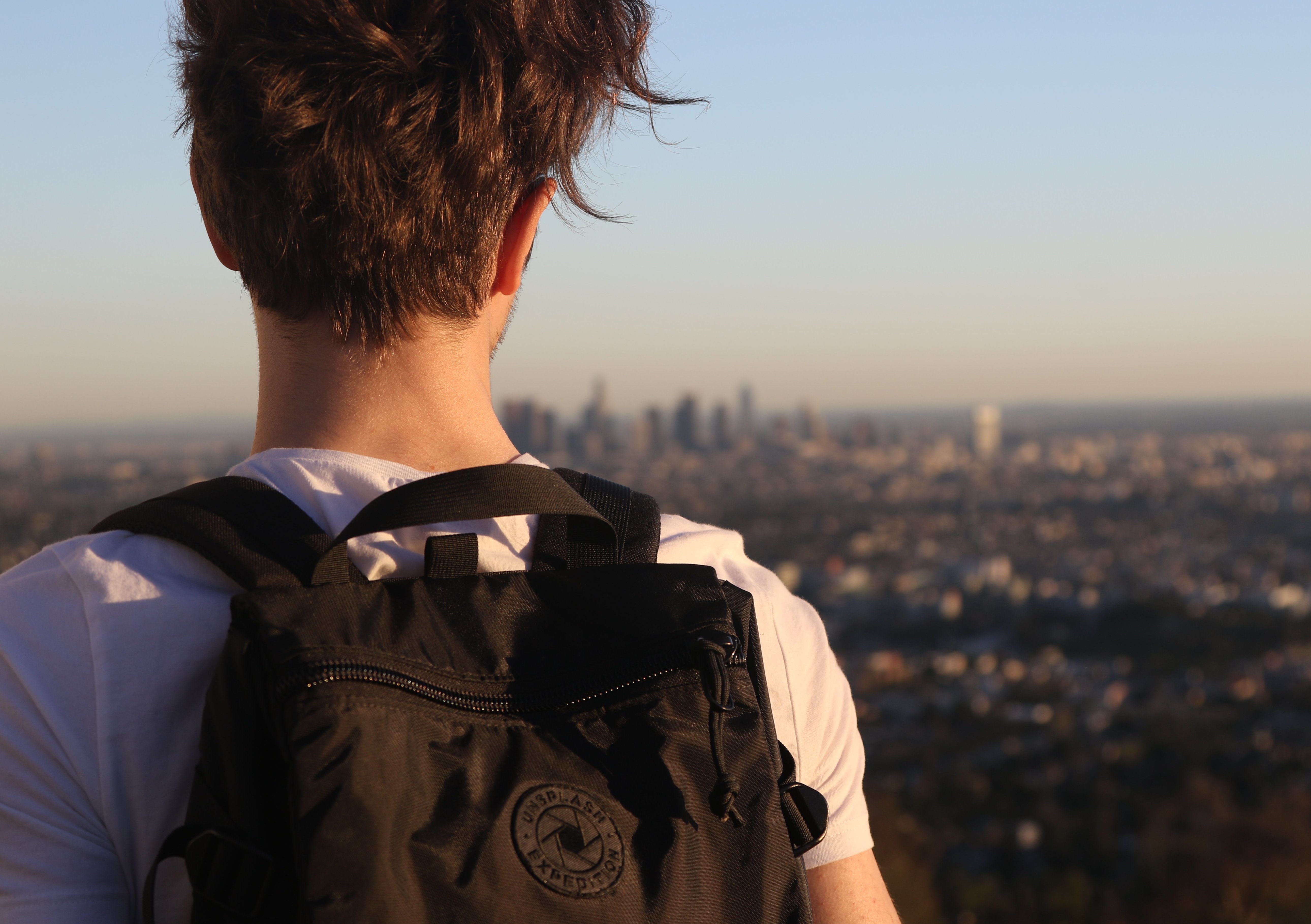 Das Bild zeigt einen jungen Mann von hinten mit einem Rucksack, der vor der Skyline von Los Angeles steht.