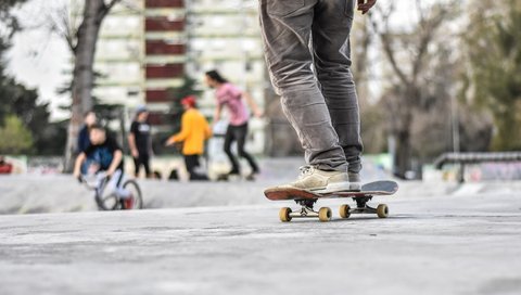 Auf dem Bild sieht man eine Person auf einem Skateboard. Im Hintergrund sind Jugendliche zu sehen. 
