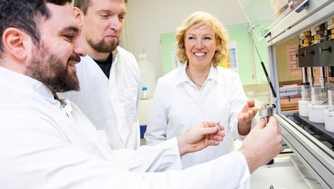 Zwei Männer und eine Frau in weißen Kitteln stehen vor einer Maschine, welche die Kaubelastung für Implantate simuliert.