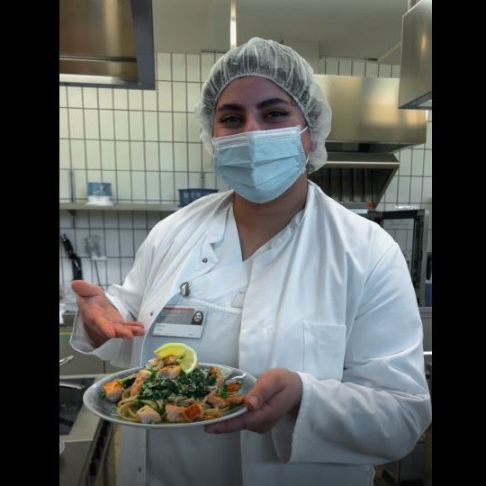 Eine Frau mit weißem Kittel und Mund-Nasen-Schutz zeigt auf einen Teller mit Lachs-Spinat-Pasta.