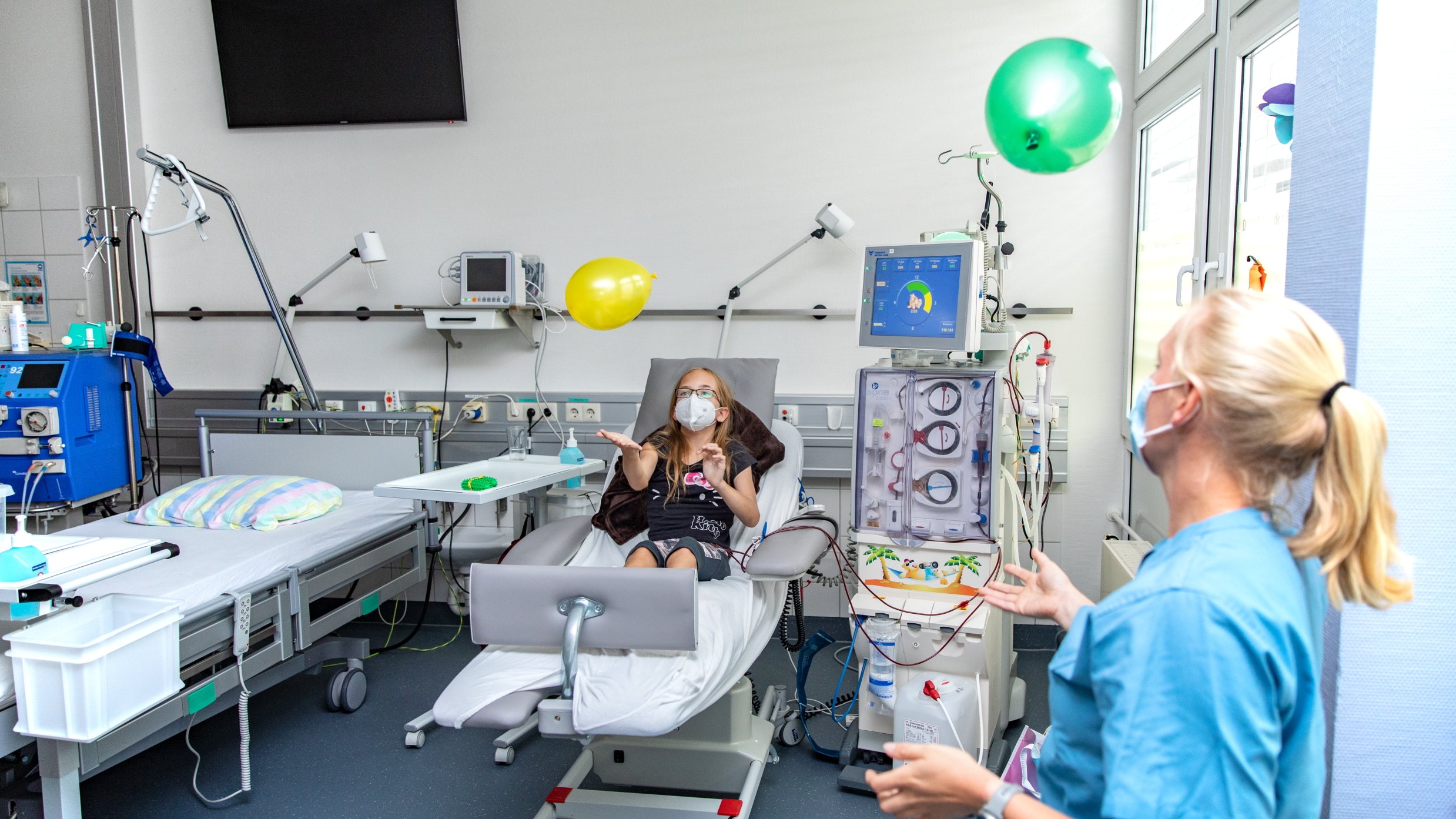 Die Patientin Jasmine sitzt im Bett und streckt sich nach Luftballons aus, Therapeutin Johanna Boyen schaut zu.