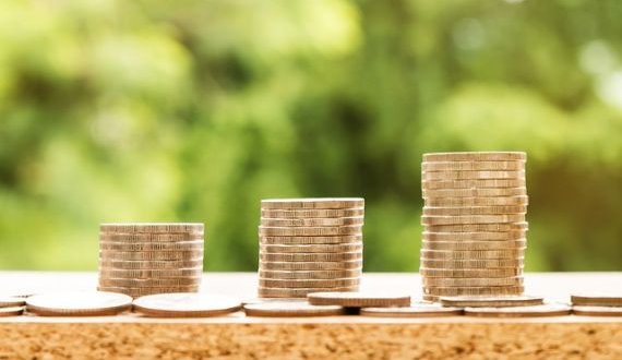 Copyright: Nattanan Kanchanaprat/Pixabay_three piles of coins on a wooden plate, all with a different height. In front of these piles lie some coins separatly. In the back some bushes can be seen.