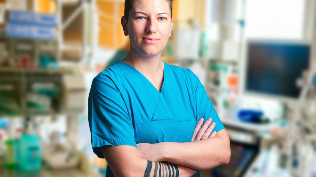 Eine Pflegekraft im blauen Kasack steht in einem Patientenzimmer mit verschiedenen intensivmedizinischen Geräten. 