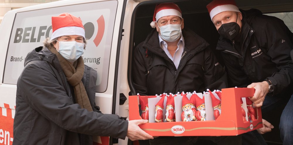 Zwei Männer stehen in der geöffneten Tür eines Lieferwagens und reichen einem dritten Mann eine kleine Palette Schokoweihnachtsmänner an. Alle drei tragen Weihanchtsmützen.