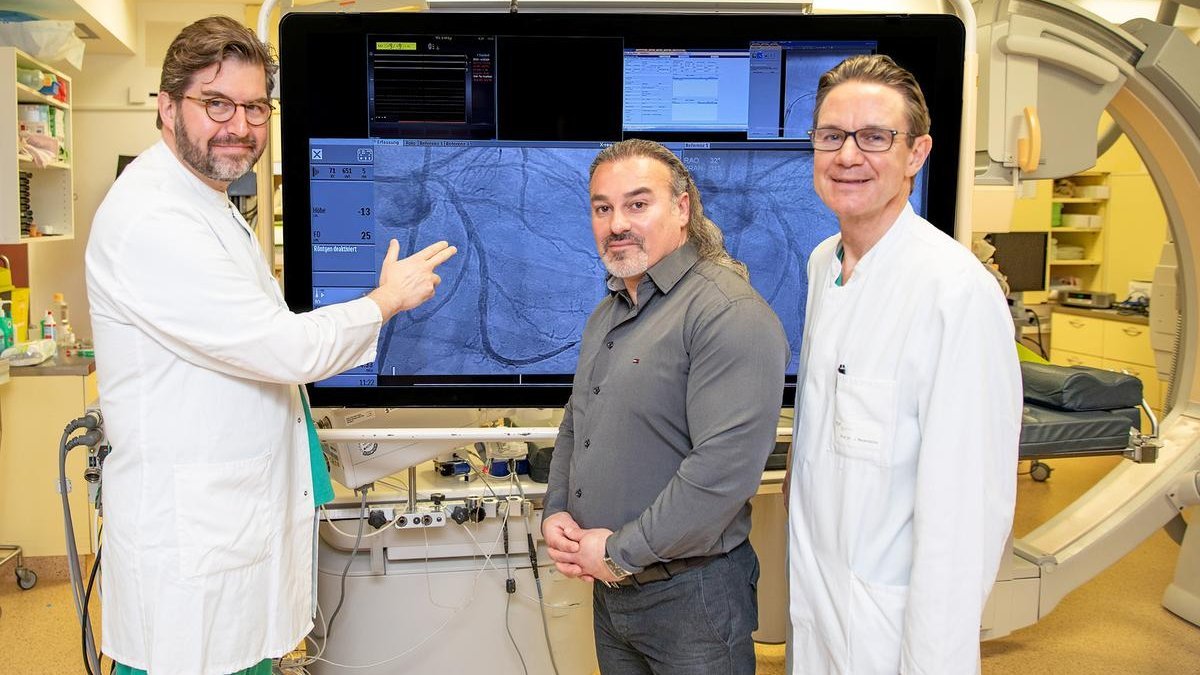 Zwei Ärzte in weißem Kittel und ein Patient stehen vor einem großen Bildschirm mit einer Aufnahme eines Herzkranzgefäßes. 