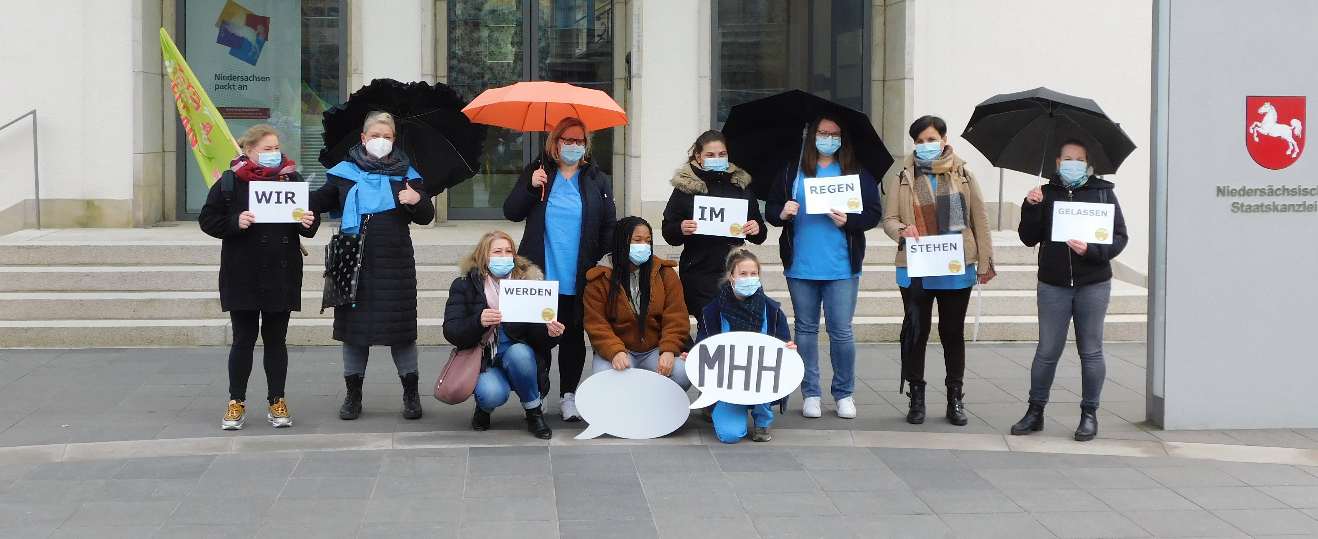 Mehrere Kolleg*innen aus der Pflege der MHH stehen mit Regenschirmen und Sprechblasen vor der Staatskanzlei in Hannover