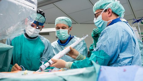 Doctors in surgical clothes stand at an intervention table and use a rod-shaped catheter system to guide the mitral valve prosthesis into a patient's heart