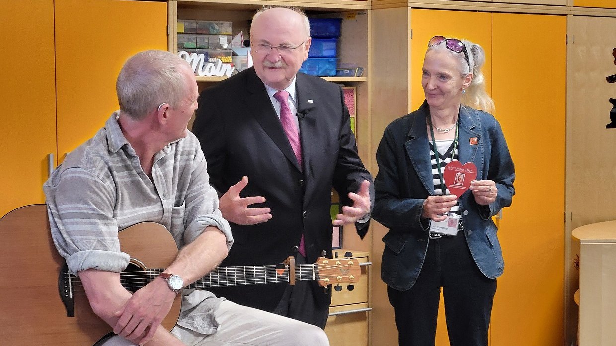 Musiker Robby Ballhause sitzt mit einer Gitarre auf einem Hocker, neben ihm stehen MHH-Präsident Prof. Manns uns Ira Thorsting vom Verein Kleine Herzen. 