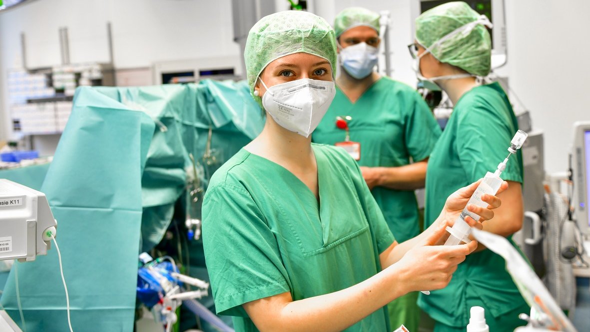 Drei Pflegekräfte in grüner OP-Schutzkleidung und mit Mund-Nasen-Schutz stehen in einem OP-Saal. Eine der Personen hält eine Spritze in den Händen. 
