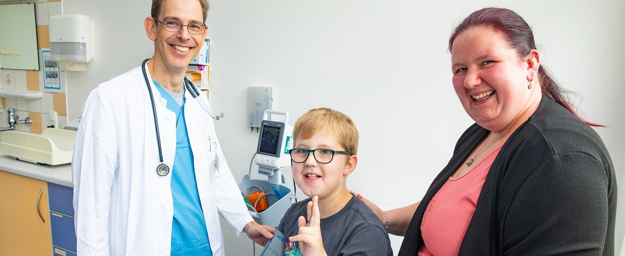 Eine Frau und ein Arzt stehen an einem Patientenbett der MHH, auf dem ein neunjähriger Junge sitzt und in die Kamera winkt. 
